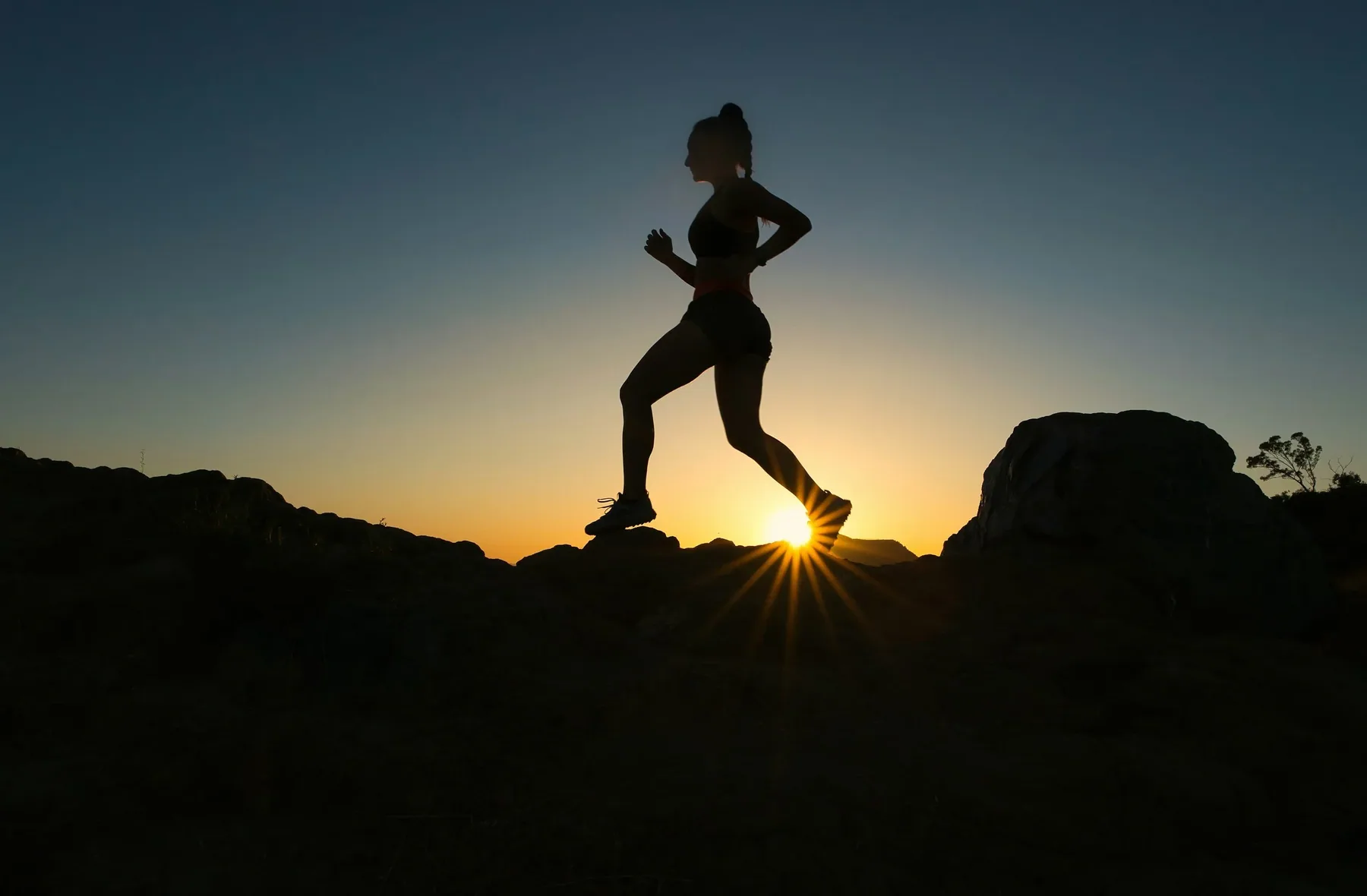 Silhouette d'une femme courant au coucher du soleil sur un terrain rocheux.